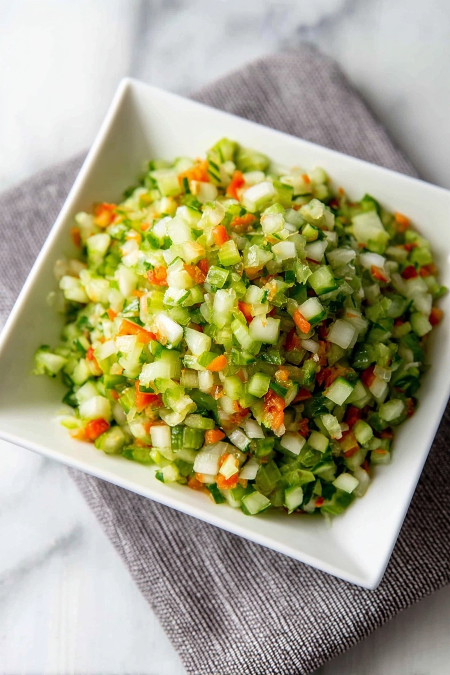 Dill Pickle Salsa, dill pickle salsa recipe, easy pickle salsa, tangy salsa with pickles, quick dill salsa - A close-up view of finely chopped vegetables in a white square bowl placed on a grey cloth on a white marbled surface. The mix has small pieces with mostly green colors from celery or cucumber, bright orange bits from carrots, and some white pieces from onions, creating a fresh and colorful look. The texture appears crisp and moist, with the small vegetable pieces piled up in layers that give a sense of volume and freshness. photo taken with an iphone --ar 2:3 --v 7