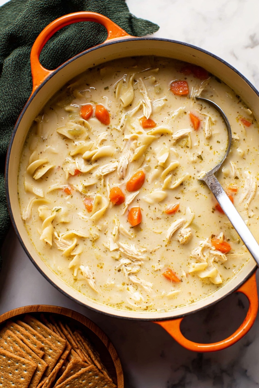 Creamy Chicken Noodle Soup, comforting chicken noodle soup, homemade chicken soup, easy creamy soup recipe, hearty chicken soup - A close-up of a white pot filled with creamy chicken noodle soup, showing a thick light beige broth mixed with small bright orange carrot slices, soft shredded white chicken pieces, and flat light yellowish noodles scattered throughout. A silver ladle is partially dipped in the soup, resting on the right side of the pot. The pot has an orange handle on each side and sits on a white marbled surface. Below the pot, there is a stack of brown crackers arranged neatly in a small wooden bowl. A dark green cloth is folded behind the pot, adding contrast to the scene. Photo taken with an iphone --ar 2:3 --v 7