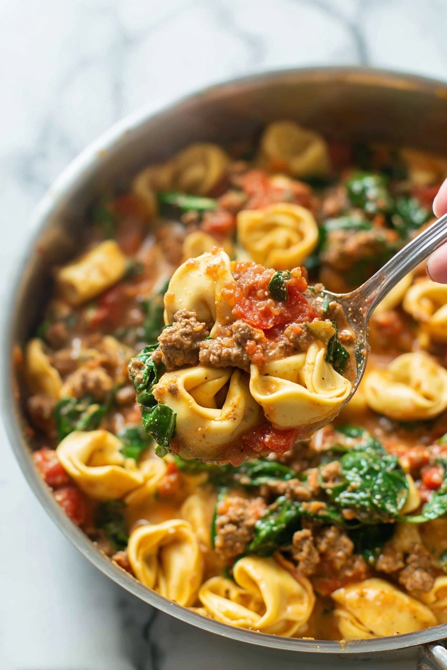 One-Pan Sausage Tortellini, sausage tortellini skillet, quick sausage pasta, creamy tortellini dinner, easy one-pan pasta - A close-up image of a cooked pasta dish in a silver pan shows three main layers. The bottom layer is pasta tortellini, small ring-shaped with light yellow color and slightly ruffled edges. The middle layer features ground meat chunks mixed with pieces of red tomato, all coated in a creamy, light brown sauce. The top layer has fresh green spinach leaves scattered throughout, adding vibrant color and texture. A woman's hand holding a silver spoon lifts a portion of the pasta, meat, tomato, and spinach mixture above the pan. The background is a white marbled surface. photo taken with an iphone --ar 2:3 --v 7