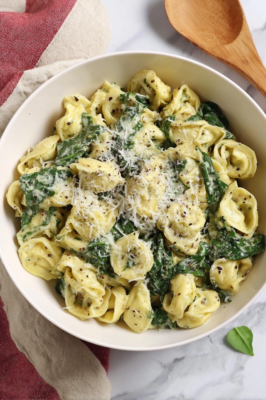 Creamy Pesto Tortellini, pesto tortellini recipe, quick pasta dinner, easy pesto pasta, creamy tortellini dinner - A white bowl filled with three layers of tortellini pasta that is light yellow, mixed with dark green spinach leaves, and coated in a creamy sauce with flecks of black pepper and herbs. On top, there is a light sprinkle of grated white cheese finely covering some pasta and spinach. The bowl sits on a white marbled surface with a beige and red cloth folded beside it, and a wooden spoon rests nearby, all softly lit in natural light. Photo taken with an iphone --ar 2:3 --v 7