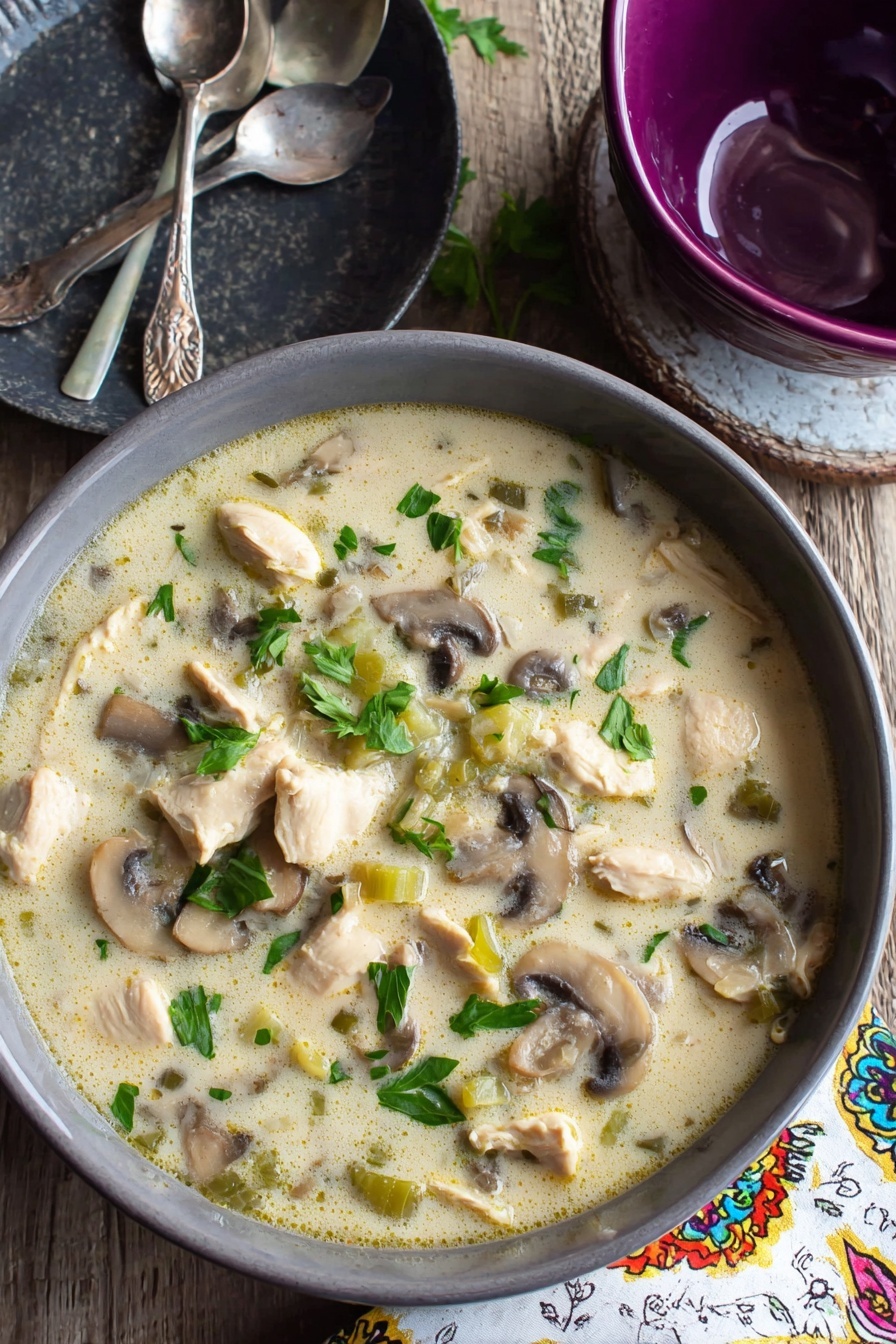 Creamy Chicken Mushroom Soup, Chicken Mushroom Soup recipe, comforting mushroom soup, easy chicken and mushroom soup, hearty creamy soup - A gray bowl filled with creamy soup that has chunks of light beige cooked chicken, sliced brown and white mushrooms, and small pieces of green celery, all mixed in a pale, creamy broth with green parsley leaves scattered on top, sitting on a wooden table next to a dark gray plate with two silver spoons and a purple bowl, with a white napkin that has colorful patterns to the side, the surface beneath is a white marbled texture photo taken with an iphone --ar 2:3 --v 7