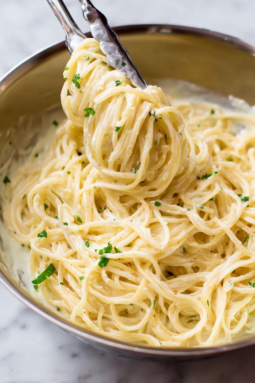 Garlic Parmesan Pasta, creamy garlic pasta, quick pasta dinner, one-pot pasta, Parmesan pasta recipe - A close-up view of a stainless steel bowl filled with creamy spaghetti pasta covered in a smooth, pale yellow sauce. The pasta strands are thick and smoothly coated, twirled around a pair of metal tongs that are lifting some of the noodles from the bowl. Small bits of green herbs are sprinkled lightly over the pasta, adding a touch of color contrast. The background is a white marbled texture. photo taken with an iphone --ar 2:3 --v 7