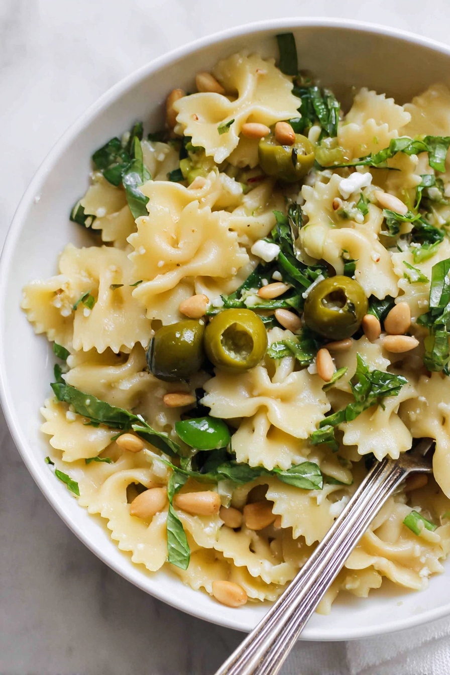 Dirty Martini Pasta Salad, olive pasta salad, blue cheese pasta salad, flavorful pasta salad, easy pasta salad recipes - A close-up view of a white bowl filled with one layer of wavy-edged pasta pieces that are light yellow in color. Scattered on top are halved green olives, fresh green leafy herbs, and a few light brown pine nuts. There are also small pieces of a creamy white ingredient and some garlic cloves. A silver fork rests inside the bowl at the bottom right corner. The bowl is placed on a white marbled surface. Photo taken with an iphone --ar 2:3 --v 7