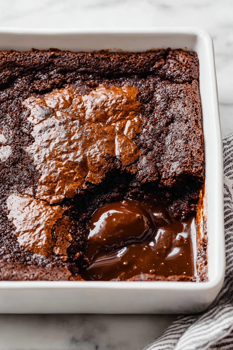 Hot Fudge Cake with Self-Saucing Chocolate, Chocolate Self-Saucing Cake, Gooey Chocolate Dessert, Easy Chocolate Cake, Warm Fudge Cake - A square white ceramic dish holds a rich chocolate dessert with a cracked, shiny top layer that is dark brown and slightly rough in texture. One section near the front corner is scooped out, revealing a thick, glossy, melted chocolate layer beneath the top crust. The melted chocolate is smooth and dark, contrasting with the more solid and matte top layer. The dish sits on a white marbled surface, and a striped cloth partially visible under one edge. photo taken with an iphone --ar 2:3 --v 7
