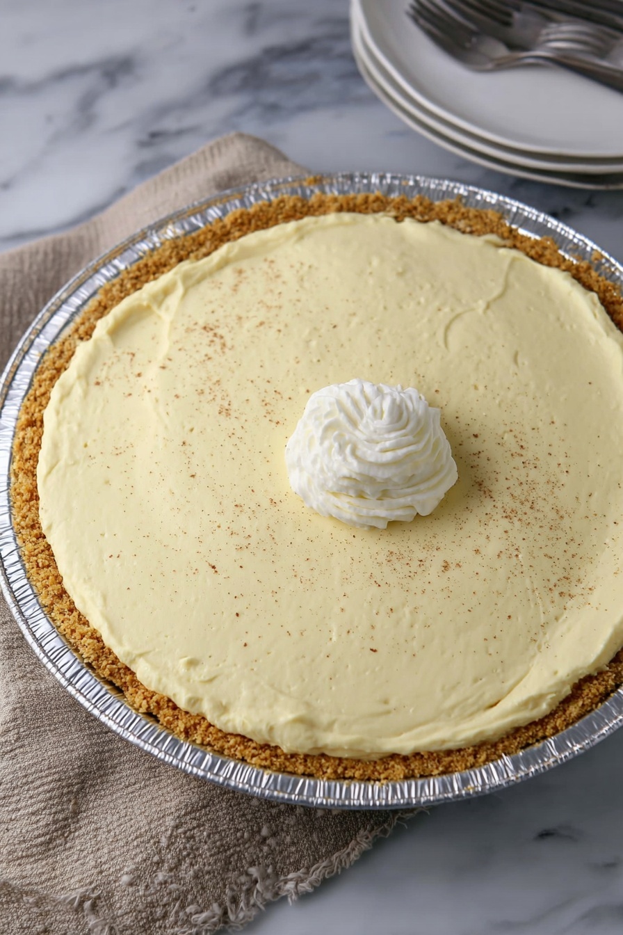 No Bake Eggnog Pie, Eggnog Pie Easy, Festive Holiday Dessert, No Bake Holiday Pie, Creamy Eggnog Pie - A creamy light yellow pie with one slice missing is in a shiny round aluminum pan. It has two layers: a thick light brown crumb crust at the bottom and a thick smooth creamy yellow filling on top. The pie is topped with a small swirl of white whipped cream in the center and sprinkled with fine brown powder all over. The pie sits on a light brown woven cloth with a white marbled surface showing at the edge. On the right side, a partial view of stacked white plates with a fork is visible. photo taken with an iphone --ar 2:3 --v 7