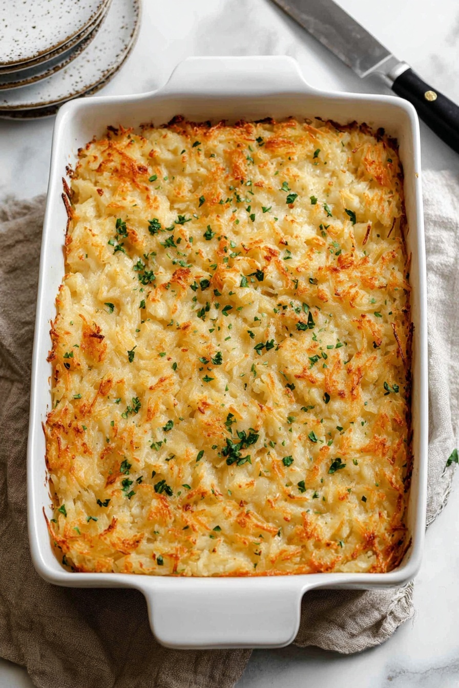 Cheddar Hashbrown Casserole, cheesy hashbrown casserole, creamy hashbrown bake, easy breakfast casserole, holiday hashbrown dish - The image shows a white rectangular baking dish filled with a golden-brown hash brown casserole topped with small green herb pieces scattered unevenly on the surface. The casserole has a crispy, textured top layer with visible shredded potatoes that have browned edges. The dish is placed on a soft beige cloth and set on a white marbled surface. Around the dish, there are white speckled side plates and a black-handled knife visible. photo taken with an iphone --ar 2:3 --v 7