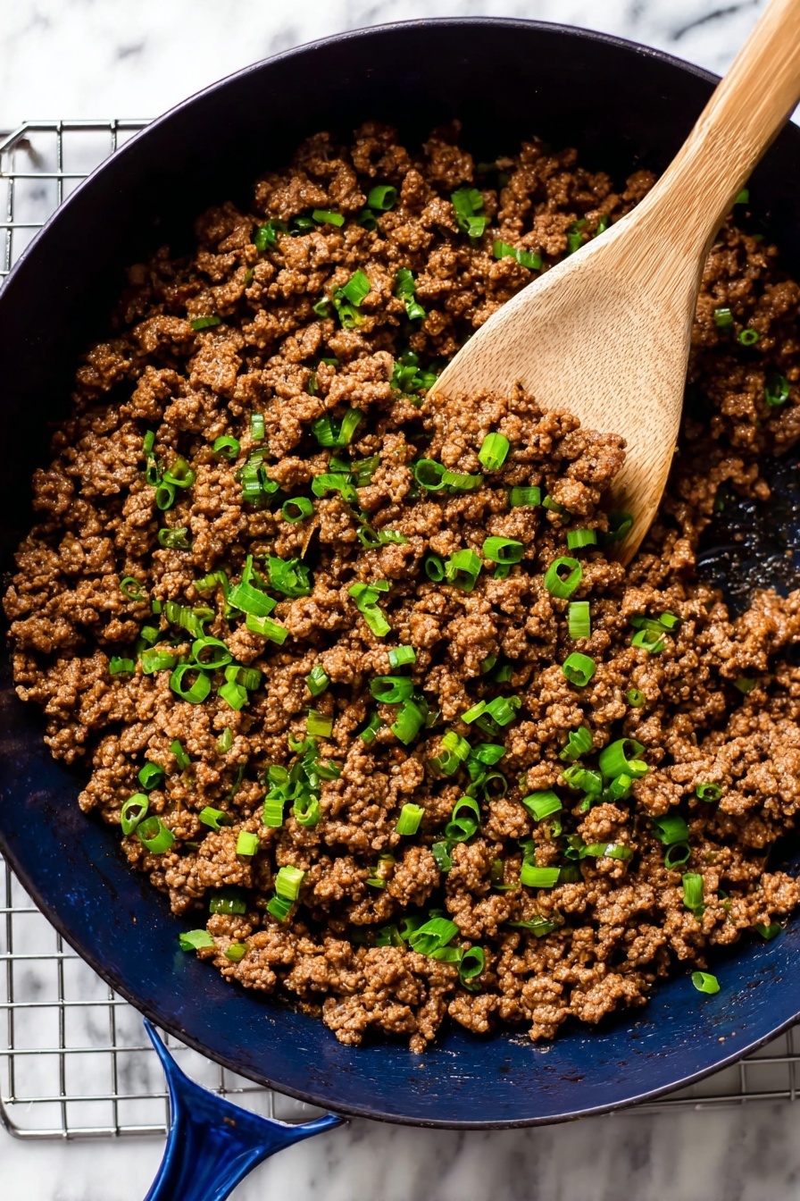 Korean Beef Bowl, Korean Beef Bowl recipe, quick Korean beef bowl, easy Korean beef bowl, flavorful beef bowl - A dark blue pan filled with cooked ground meat mixed with small pieces of green onion scattered evenly on top. The meat is rich brown with a soft texture, and the green onions add bright green spots throughout. A wooden spoon is scooping some of the meat and onions, positioned on the right side of the pan. The pan sits on a grid metal rack over a white marbled surface. Photo taken with an iphone --ar 2:3 --v 7